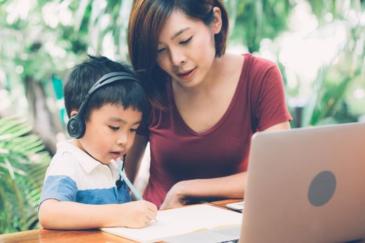 Young asian mother and son using laptop computer for study and learning together at home, boy writing on notebook for homework and wearing headphone, teacher or mom support child, education concept.