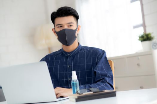 Young business man in face mask working from home with laptop computer on desk for protection for outbreak of covid-19 and healthcare, male quarantine stay home using notebook on desk, new normal.