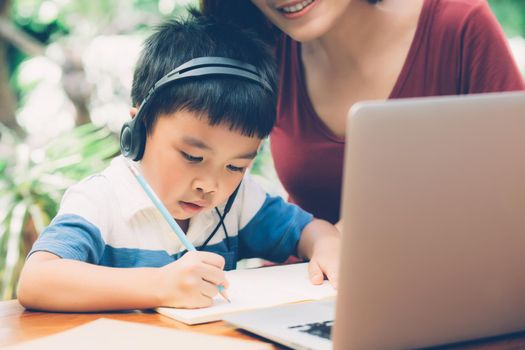 Young asian mother and son using laptop computer for study and learning together at home, boy writing on notebook for homework and wearing headphone, teacher or mom support child, education concept.