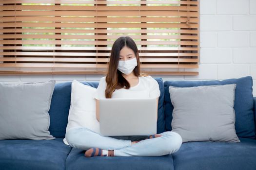 Young asian business woman in face mask work from home on laptop computer online to internet in living room, girl in medical mask and quarantine for pandemic of covid-19, stay home, new normal.