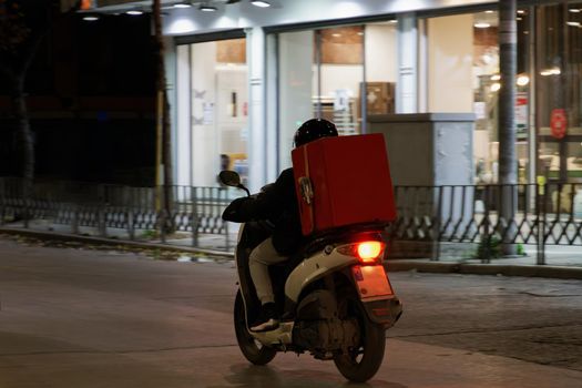 Illuminated view of male with helmet riding motorcycle to deliver take away food contained on a red box in Thessaloniki, Greece.