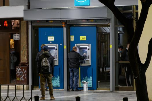 Illuminated night view of unidentified people using Hellenic 24-hour cash machines outside a branch of National Bank.