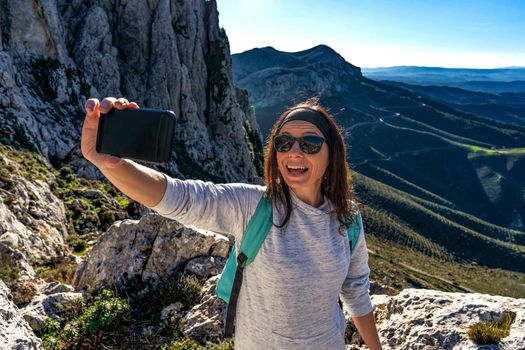 Caucasian young woman making self portrait with smartphone at open mouth on a mountain top with beautiful background panorama - Solo female traveler using internet technology to share her nature trip