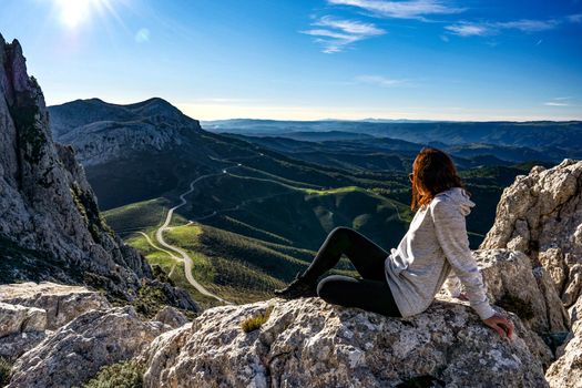 Beautiful young Caucasian woman sitting on a big white rock looking down to gorgeous panorama with a road that climbs through the mountains to the horizon - Carefree solo female traveler resting
