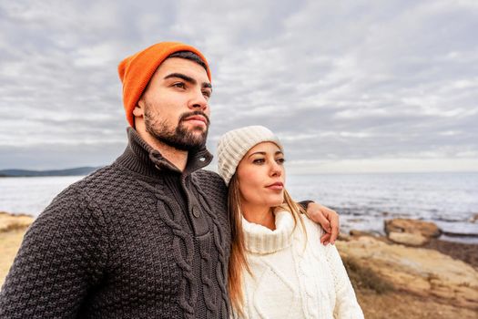 Handsome guy embracing his girlfriend outdoor on a sea resort during winter vacation watching horizon at sunset wearing sweater and wool cap for cold outdoors - Beautiful Caucasian heterosexual couple