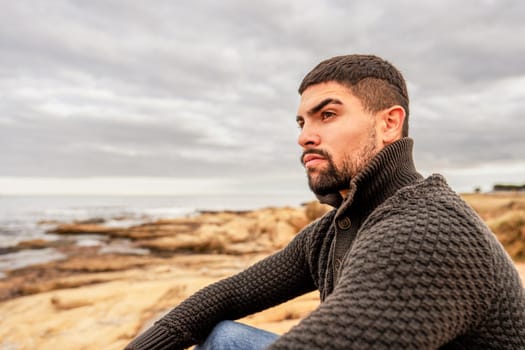 Handsome Caucasian serious young man sitting on sea rocks at sea resort watching horizon wearing sweater - Stylish guy with pensive glance with cloudy sky and orange photo filter for fall-winter mood
