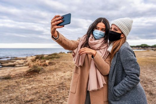 Young mixed race female LGBTQ couple making selfie with smartphone in sea resort in safe protection mask - Hispanic beautiful woman using tech to sharing photo with her lesbian girlfriend on social