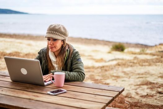 Internet female freelance working outdoor in sea resort sitting at wooden table wearing fall-winter clothes and wool hat with tea cup close to her aluminum laptop - Change your life making online job