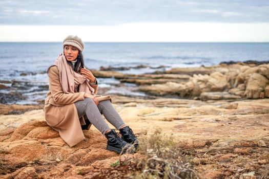 Beautiful young stylish Hispanic model sitting on a sea rock wearing fall-winter clothes, coat and wool cap in natural colors touching hair on the scarf - Glamour shot of cute woman posing outdoor