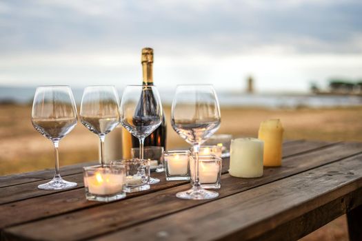 All is ready for the end of year toast in winter holidays by the sea - Celebration scene with selective focus on empty glasses, closed champagne bottle, and candles on a wooden table of outdoor bar