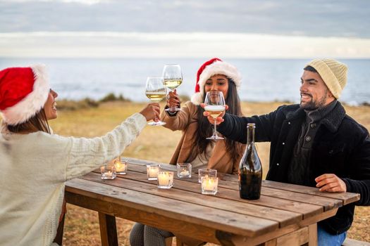 Group of young mixed race friends toasting outdoor sitting at a wooden table near the water of the ocean in sea winter holidays - Multiracial people celebrating in the nature Christmas and New Year