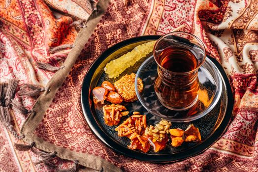 Black tea in armudu glass with candies with different nuts and crystal sugar on metal tray over tablecloth