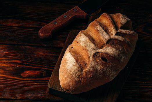 Rye loaf on cutting board with knife over dark wooden background.