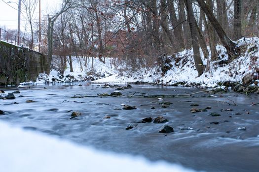 A Long Exposure of a Small Flowing Creek Next to a Snow Covered Bank With Trees and Foliage