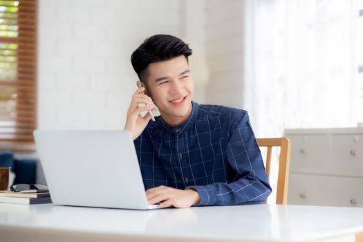 Young asian man talking phone and work from home with laptop computer, freelance using notebook to internet online and speak on smartphone, domestic life, business and communication concept.