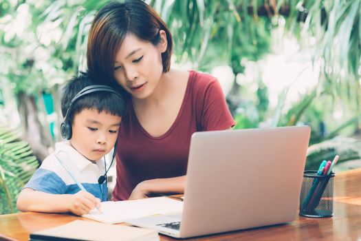 Young asian mother and son using laptop computer for study and learning together at home, boy writing on notebook for homework and wearing headphone, teacher or mom support child, education concept.