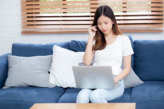 Young asian business woman smile and work from home with laptop computer online to internet on sofa in living room, freelance girl using notebook on couch with comfort, new normal, lifestyle concept.