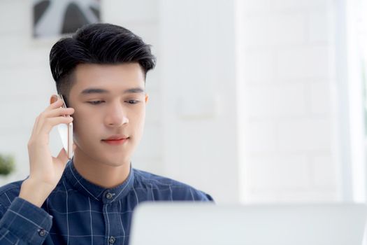 Young asian man talking phone and work from home with laptop computer, freelance using notebook to internet online and speak on smartphone, domestic life, business and communication concept.