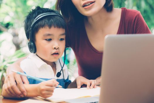 Young asian mother and son using laptop computer for study and learning together at home, boy writing on notebook for homework and wearing headphone, teacher or mom support child, education concept.