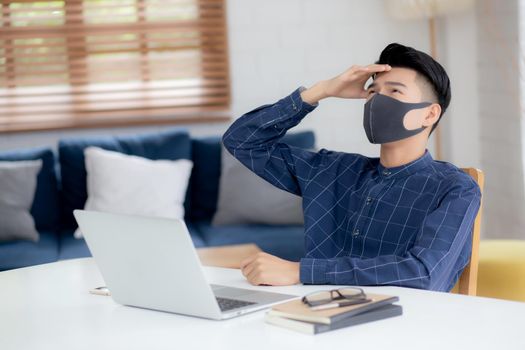 Young business man in face mask working from home with laptop computer and bored on desk for protection for outbreak of covid-19, male quarantine stay home using notebook with unhappy, new normal.