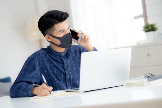 Young asian business man in face mask talking smartphone and work on laptop computer for protect covid-19, businessman quarantine work from home and writing notebook, social distancing and stay home.