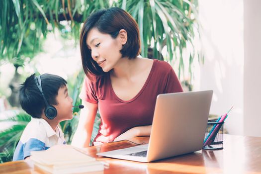 Young asian mother and son using laptop computer for study and learning together at home, boy wearing headphone for e-learning with distancing, teacher or mom support child, education concept.