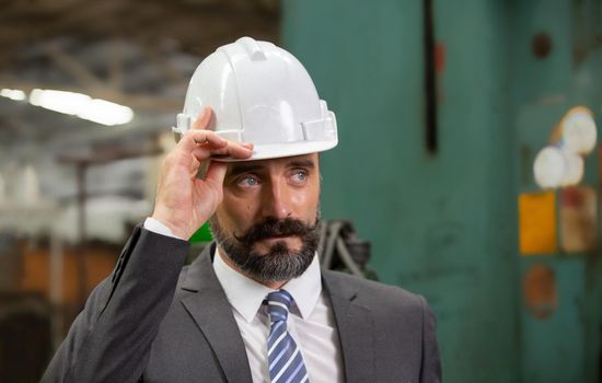 The bearded hipster director with engineer hat standing against machine in production in factory