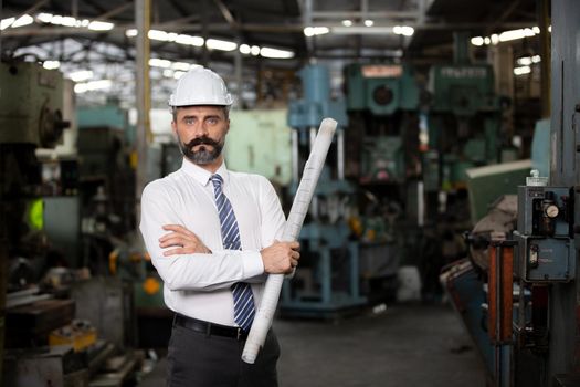 The bearded hipster director with engineer hat standing against machine in production in factory