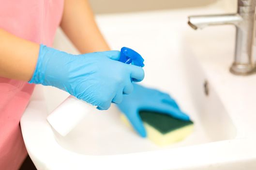 Close-up women hand in a blue rubber glove and cleaning spong in the picture, removes and washes bathroom sink and mirror.