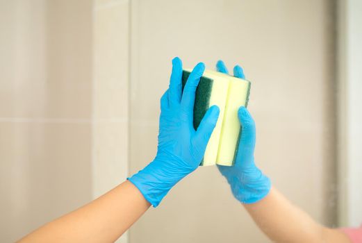 Close-up women hand in a blue rubber glove and cleaning spong in the picture, removes and washes bathroom sink and mirror.