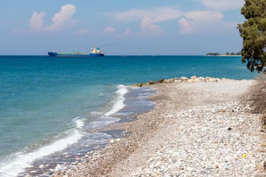 Gravel, pebble beach of Soroni Beach at Rhodes island with ocean waves, turquoise water and cargo ship in the background 