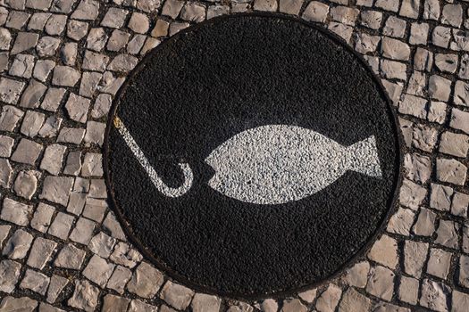A picture of a fish and a hook painted white on a black background. A view from above of the cobbled sidewalk along the Tagus River in Lisbon, Portugal. The signal informs anglers that fishing is allowed along the river bank with this notice. The image also lends itself to the concept that appearances can be deceiving.