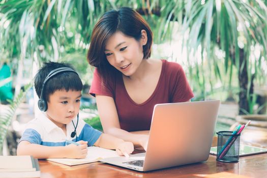Young asian mother and son using laptop computer for study and learning together at home, boy writing on notebook for homework and wearing headphone, teacher or mom support child, education concept.