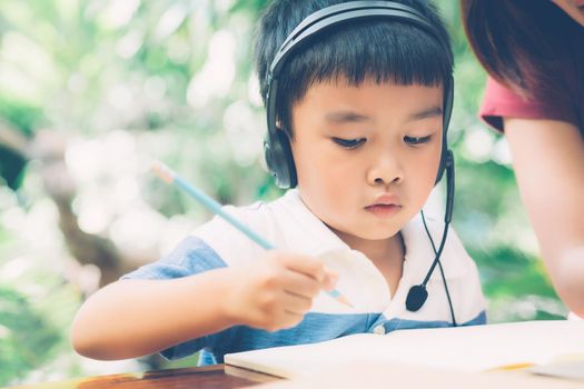 Young asian mother and son using laptop computer for study and learning together at home, boy writing on notebook for homework and wearing headphone, teacher or mom support child, education concept.