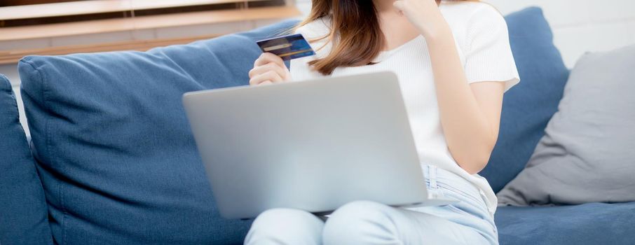 Young asian woman sit on sofa using laptop computer shopping online with credit card buying to internet, happy girl payment with e-business on couch, purchase and payment, business concept.