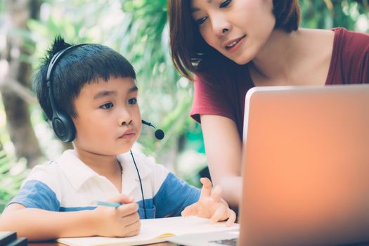 Young asian mother and son using laptop computer for study and learning together at home, boy writing on notebook for homework and wearing headphone, teacher or mom support child, education concept.