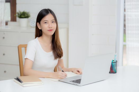 Young asian woman writing on notebook for planning working and using laptop computer on desk at home, girl notes about finance, female study and learning, business and communication concept.