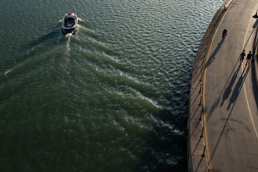 Light and shadows: a small fishing boat returns to the port-canal of the city of Pescara and  some people travel the road on the riverbank, at sunset. Horizontal composition, copy space for text.