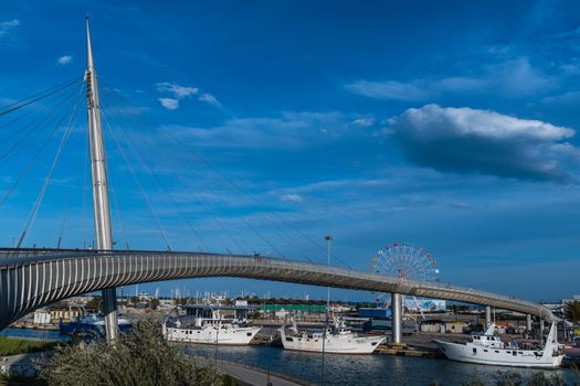 Panoramic view of the Ponte del Mare, a cycle-pedestrian bridge which connects the south coast with the north of the river Pescara. Fishing boats docked in the canal and a ferris wheel in the background. Pescara, Abruzzo, Italy.