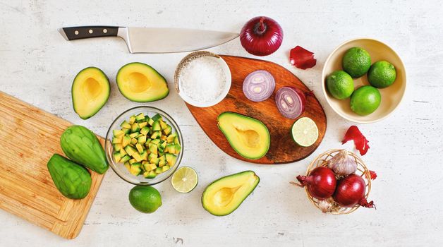 Avocado halves, pieces, limes and onions, cup with salt - basic guacamole ingredients and chef knife on white working board, flat lay photo.