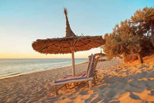 Two empty wooden sunbeds facing the sunset, sea in background, straw sun shade above, image illustrating tropical vacation.