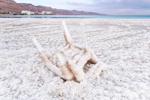 Small plastic chair completely covered with crystalline salt on shore of dead sea, closeup detail, blurred hotel resorts in distance - Ein Bokek, Israel.