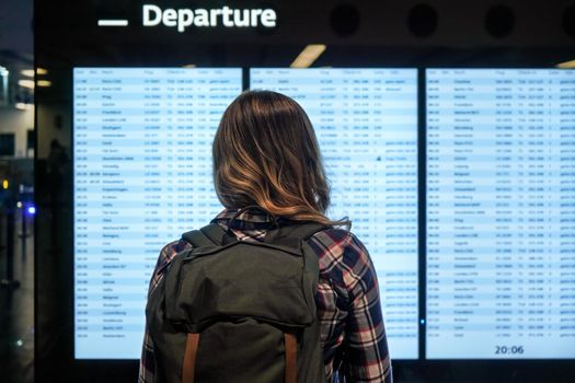 Young woman with green backpack and shirt looking at departure board screen at the airport, view from behind.