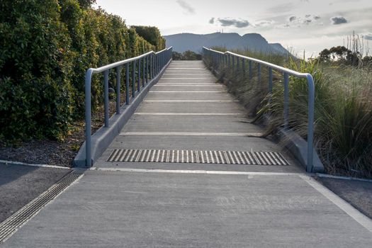 A concrete walkway with grey steel handrails leading to a carpark with hills in the background