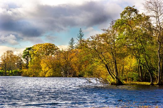 A view of the side of a lake, surrounded by trees, during autumn, with green, yellow and orange leaves, and a partly cloudy sky.