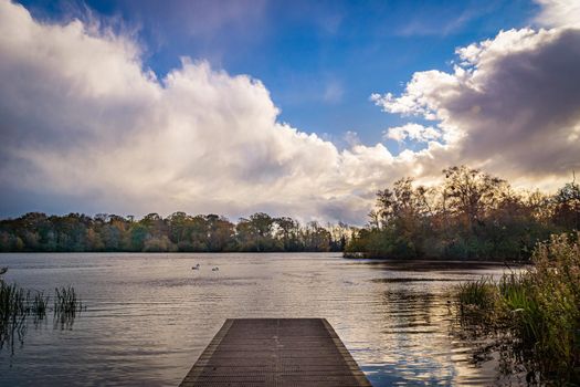 A view of a jetty on the side of a lake, surrounded by trees, during autumn, with green, yellow and orange leaves, and a cloudy sky.