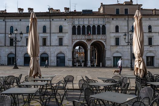 Deserted tables and chairs outside a restaurant. Only two persons walking across the main town square in Brescia during the coronavirus emergency lockdown.