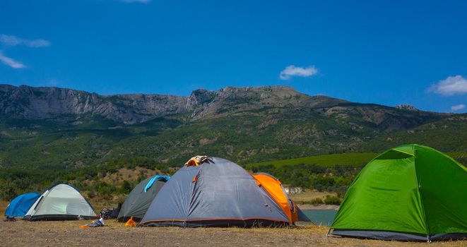 Multicolored tents on the shore of a mountain lake against the backdrop of inaccessible rocks surrounded by a green forest.