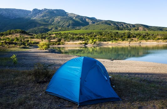 A blue tourist tent on the shore of a mountain lake against the backdrop of inaccessible rocks surrounded by a green forest.