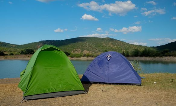 Multicolored tents on the shore of a mountain lake against the backdrop of inaccessible rocks surrounded by a green forest.